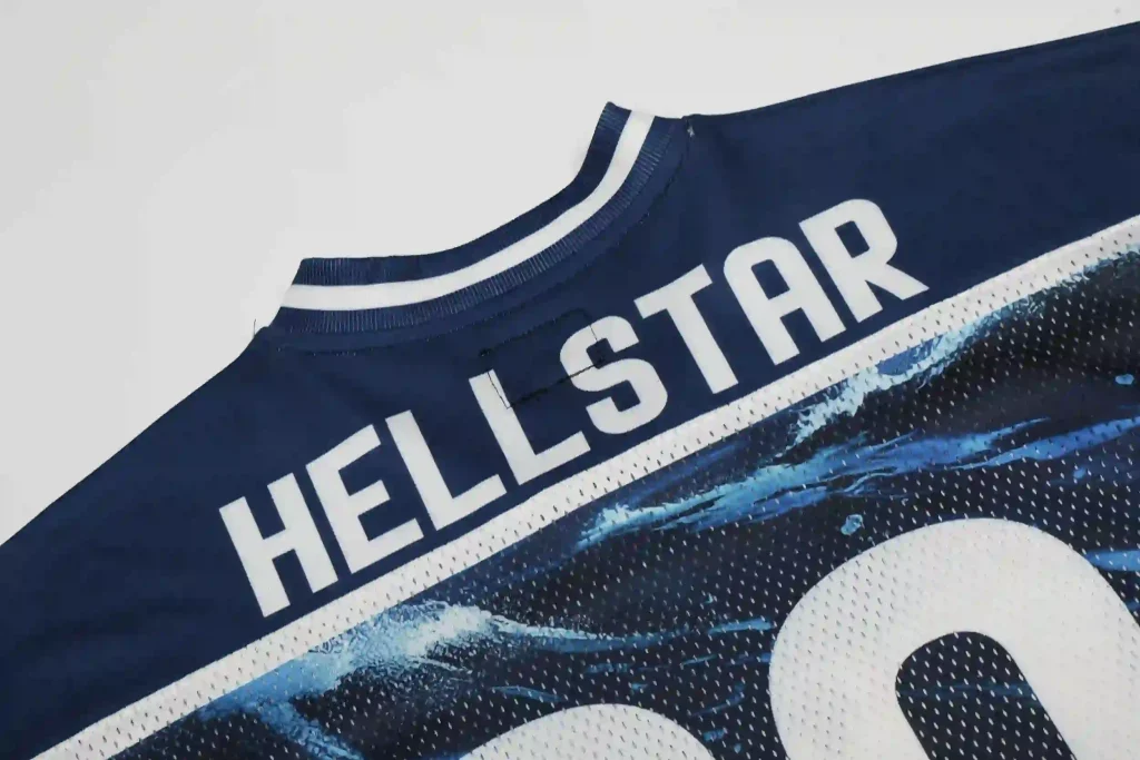 Close-up of a navy blue Hellstar mesh jersey-style top, featuring bold white "HELLSTAR" lettering across the chest, white ribbed trim on the crew neck, and a blue wave graphic print on the perforated mesh fabric